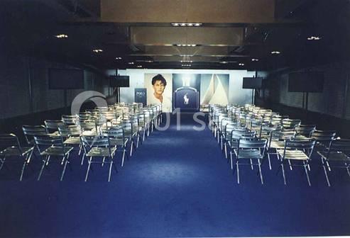 Salle de conférence avec des rangées de chaises métalliques alignées sur un tapis bleu, affiches publicitaires au fond.