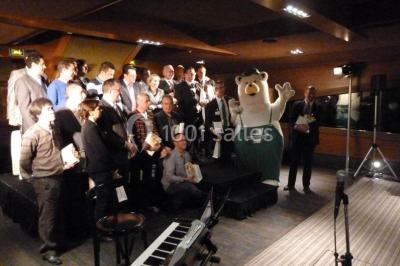 Un groupe de personnes pose sur une scène éclairée, accompagné d'une mascotte en costume d'ours.