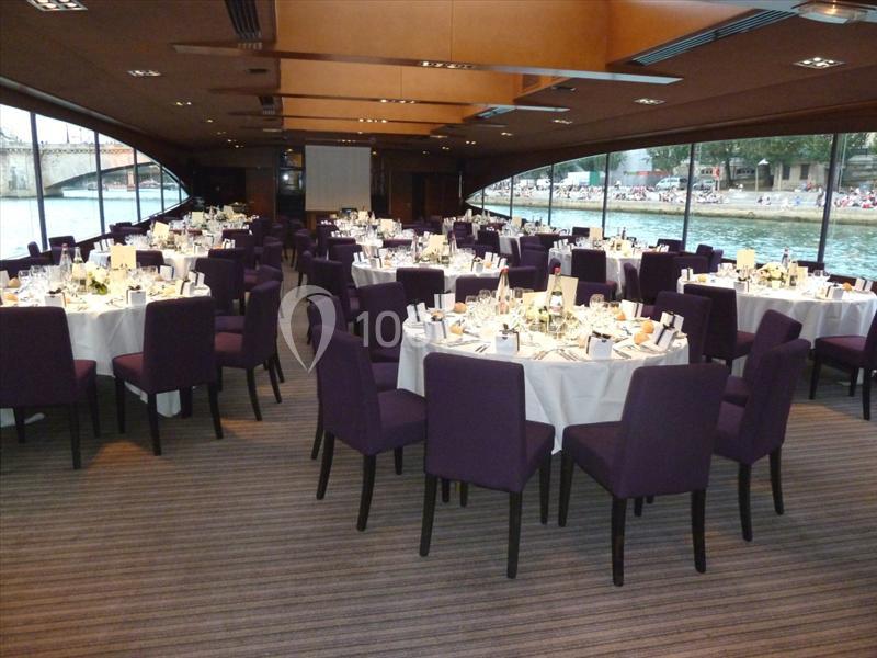 Salle de réception avec des tables rondes dressées, chaises violettes et vue sur un paysage fluvial à travers de grandes…