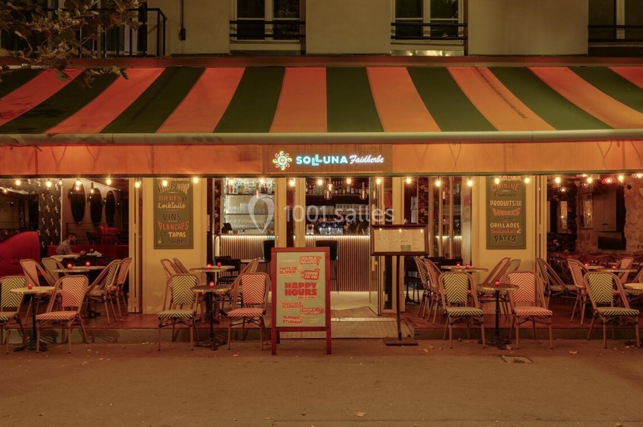 Façade d'un restaurant avec terrasse éclairée, chaises en osier et auvent rayé orange et vert, en soirée.