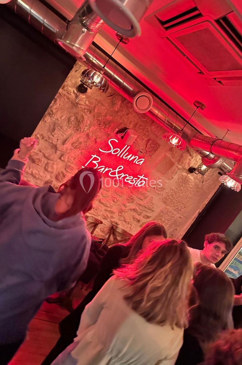 Des personnes discutent et dansent dans un bar à l'éclairage rouge, avec un mur en pierre et un néon ’Solluna Bar & Resto’.