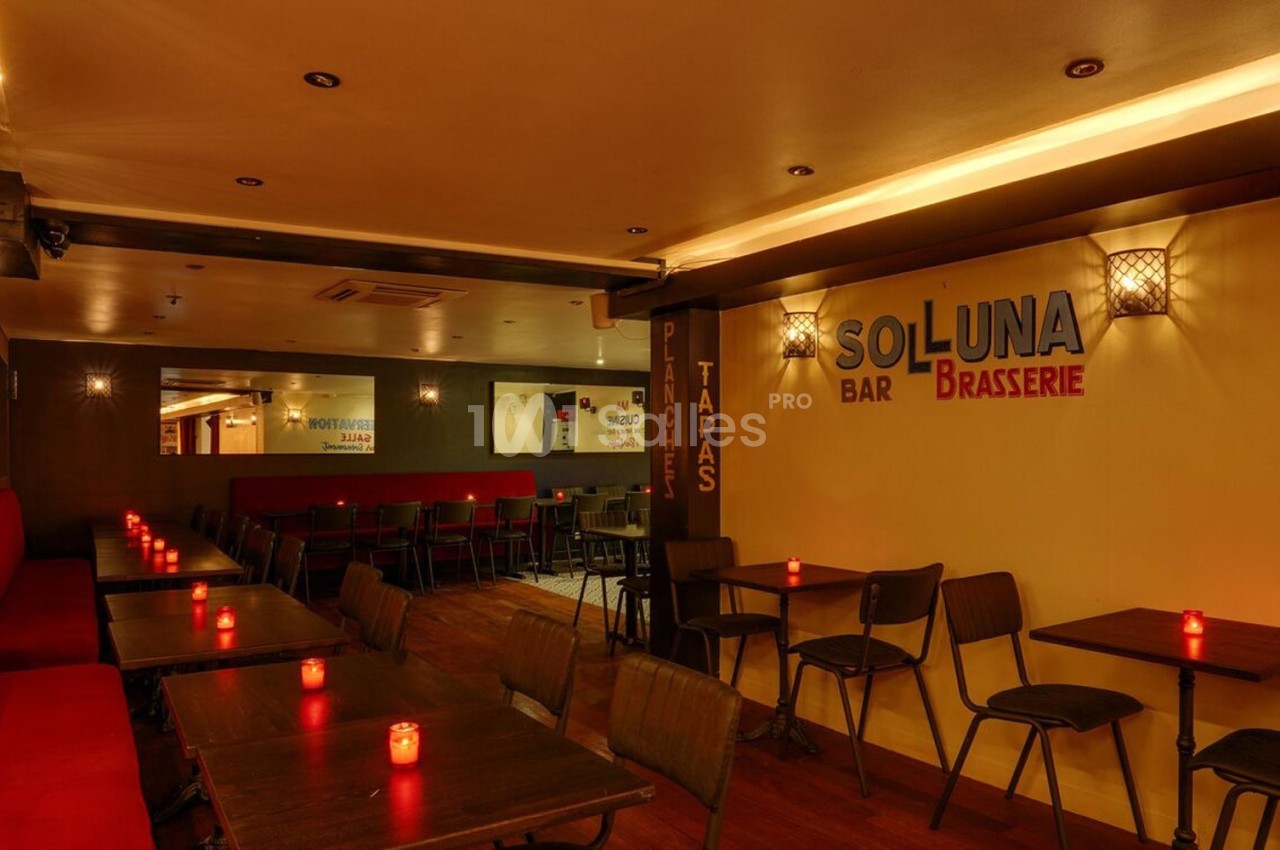 Salle de restaurant chaleureuse avec éclairage tamisé, tables en bois, chaises noires et bougies rouges allumées.