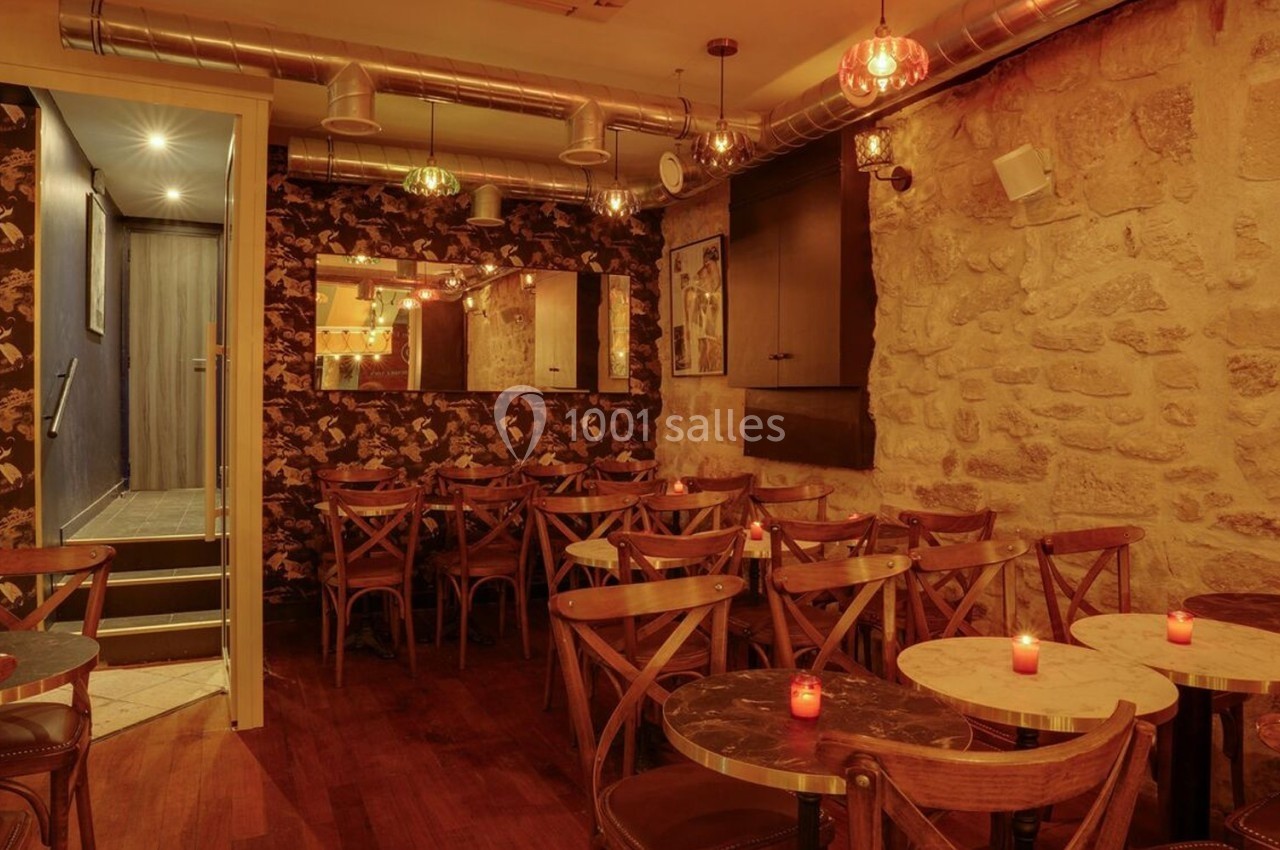 Salle de restaurant chaleureuse avec murs en pierre, tables rondes, chaises en bois et bougies allumées.