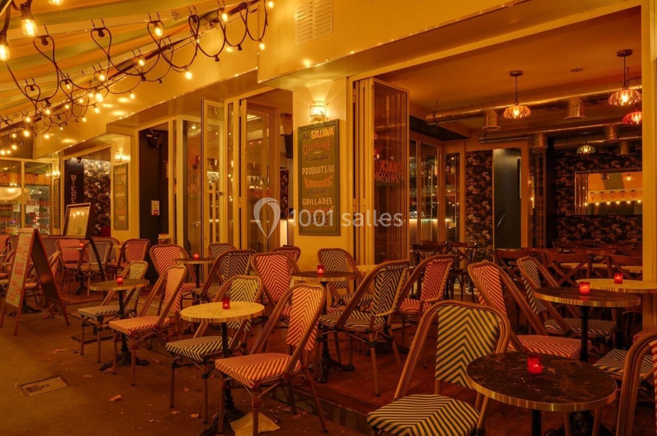 Terrasse d'un café-restaurant éclairée par des guirlandes lumineuses, avec des tables et chaises colorées.