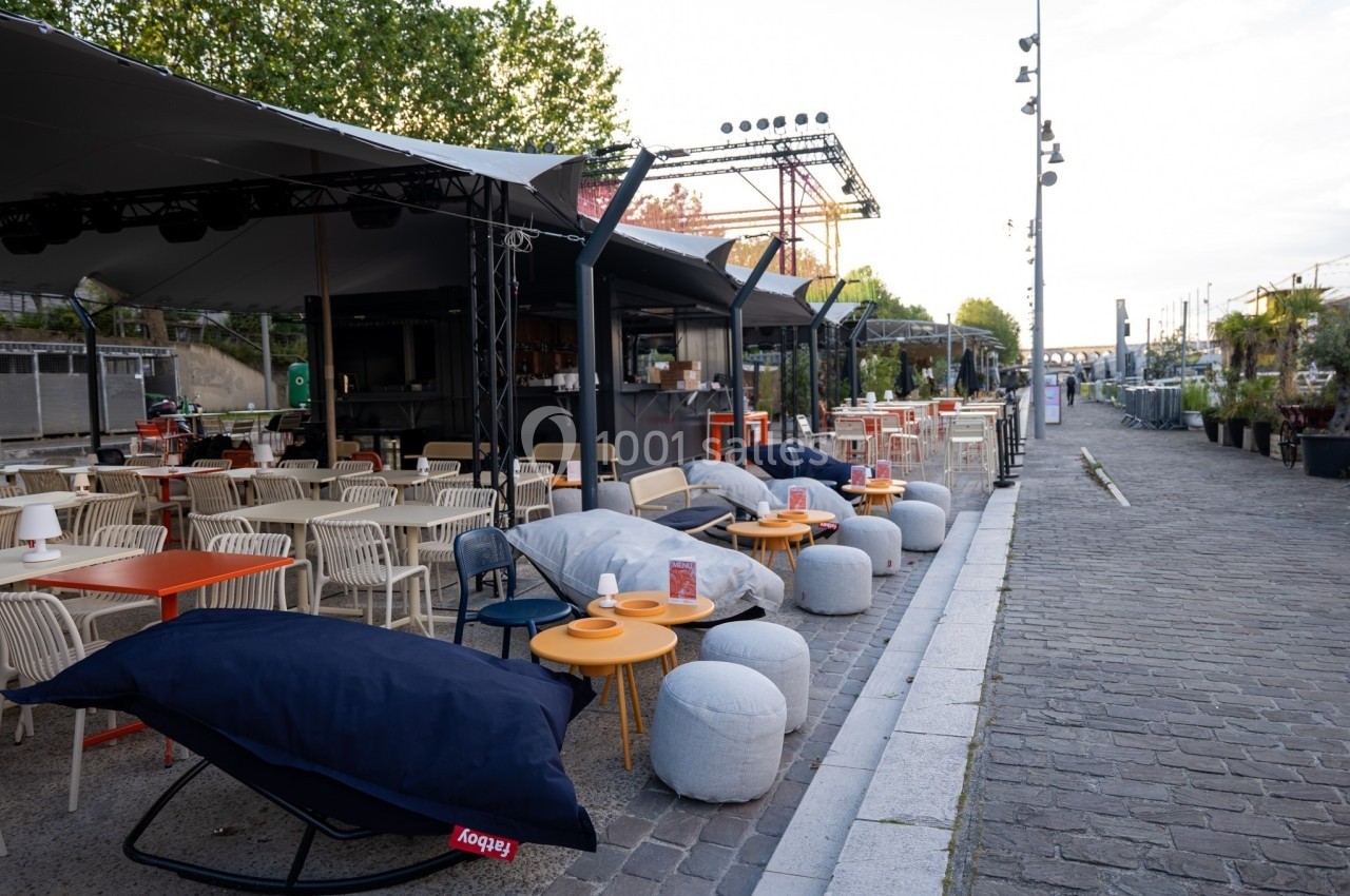Terrasse extérieure avec tables, chaises et poufs colorés, située le long d'une promenade pavée près d'un fleuve.