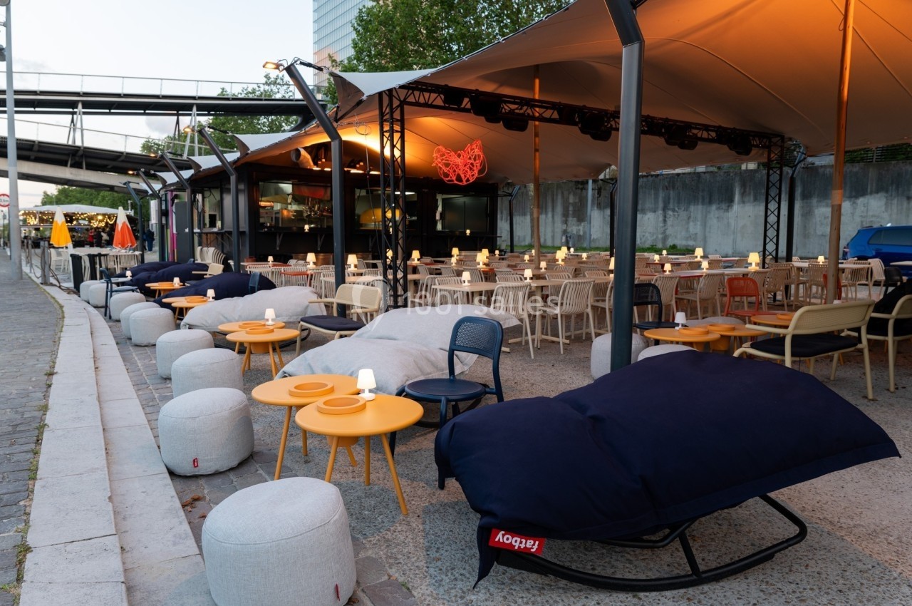 Terrasse extérieure avec mobilier coloré, tables éclairées et poufs, sous une structure couverte près d'un quai.