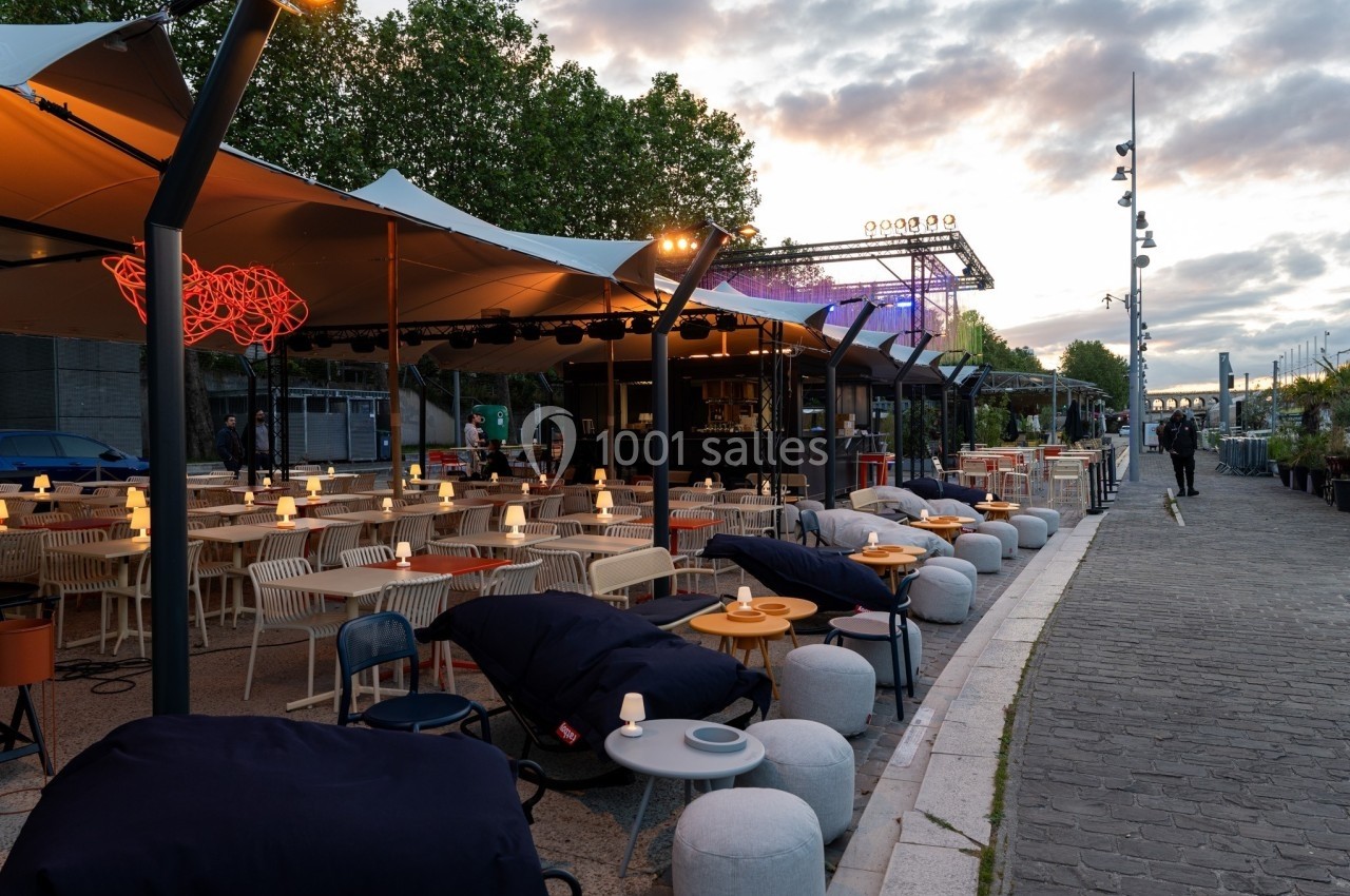 Terrasse extérieure avec tables, chaises et éclairage tamisé au bord d'une promenade, sous un ciel nuageux au crépuscule.
