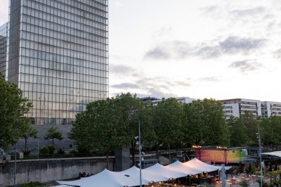 Vue d'un quai animé avec des stands colorés sous des tentes, près d'un bâtiment moderne et d'un cours d'eau.