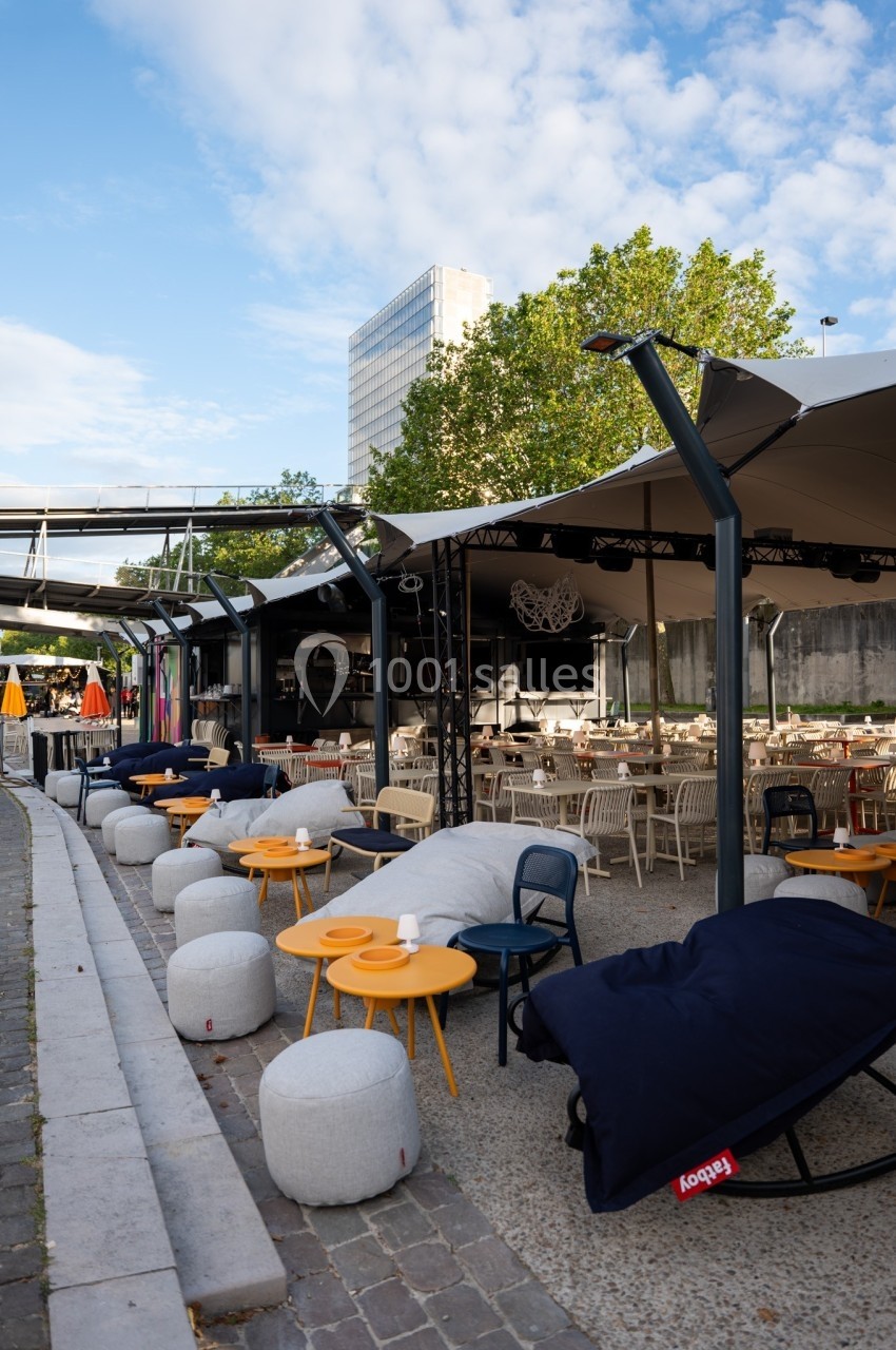 Terrasse extérieure avec tables, chaises, poufs et parasols, située près d'un bâtiment moderne et d'arbres.