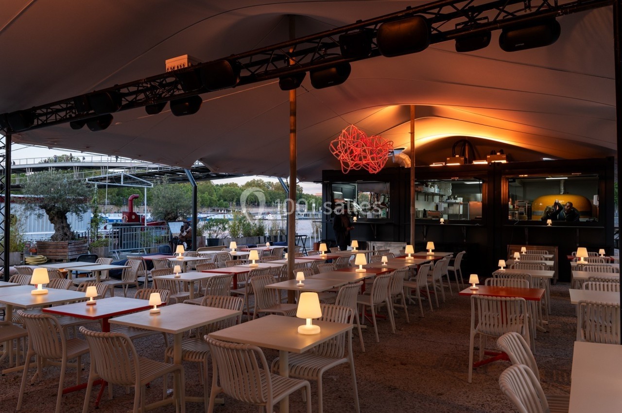 Terrasse de restaurant en soirée avec tables éclairées par des lampes, vue sur un fleuve et cuisine ouverte en arrière-plan.