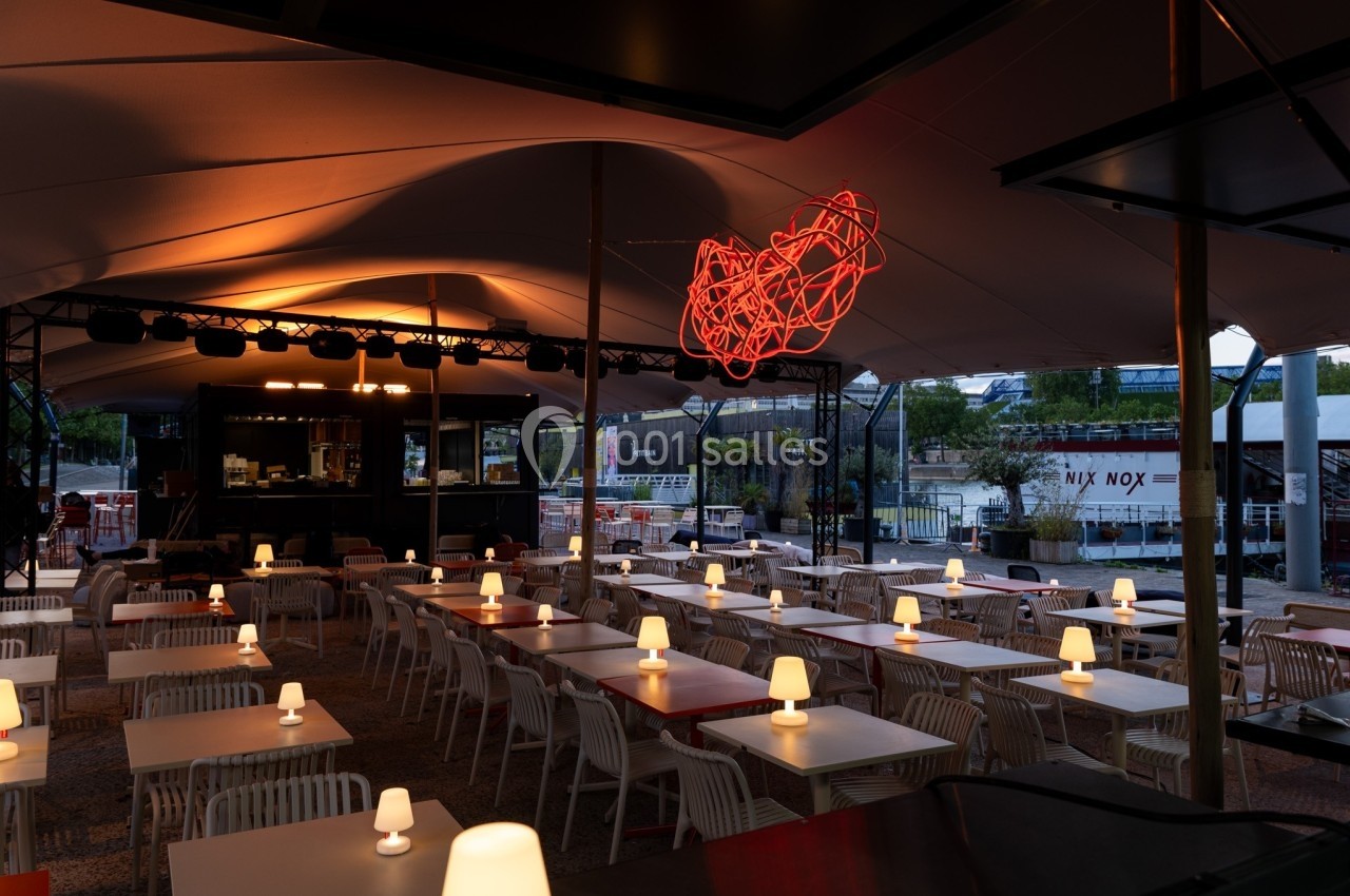 Terrasse couverte avec tables et chaises blanches, éclairée par des lampes de table et une sculpture lumineuse rouge.