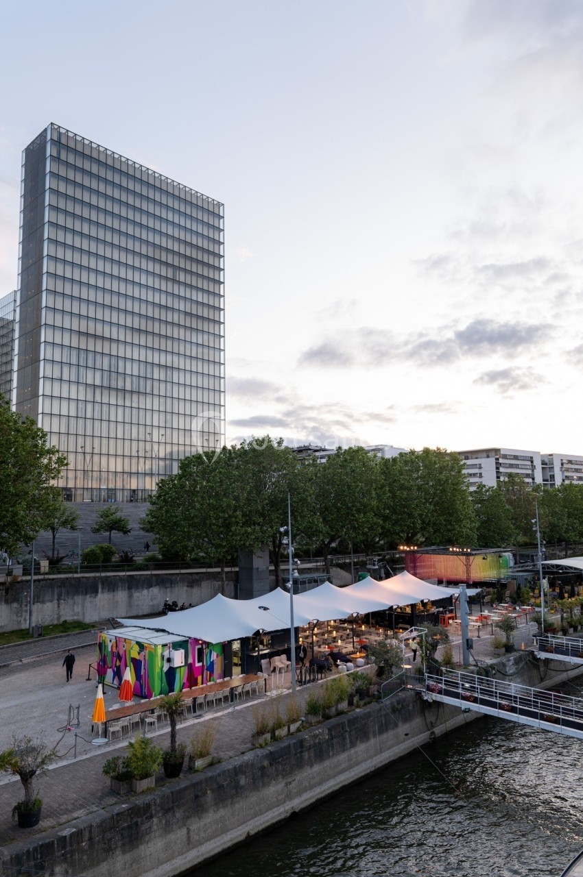 Vue d'un marché en plein air au bord de la Seine, avec des stands colorés et un grand immeuble en arrière-plan.