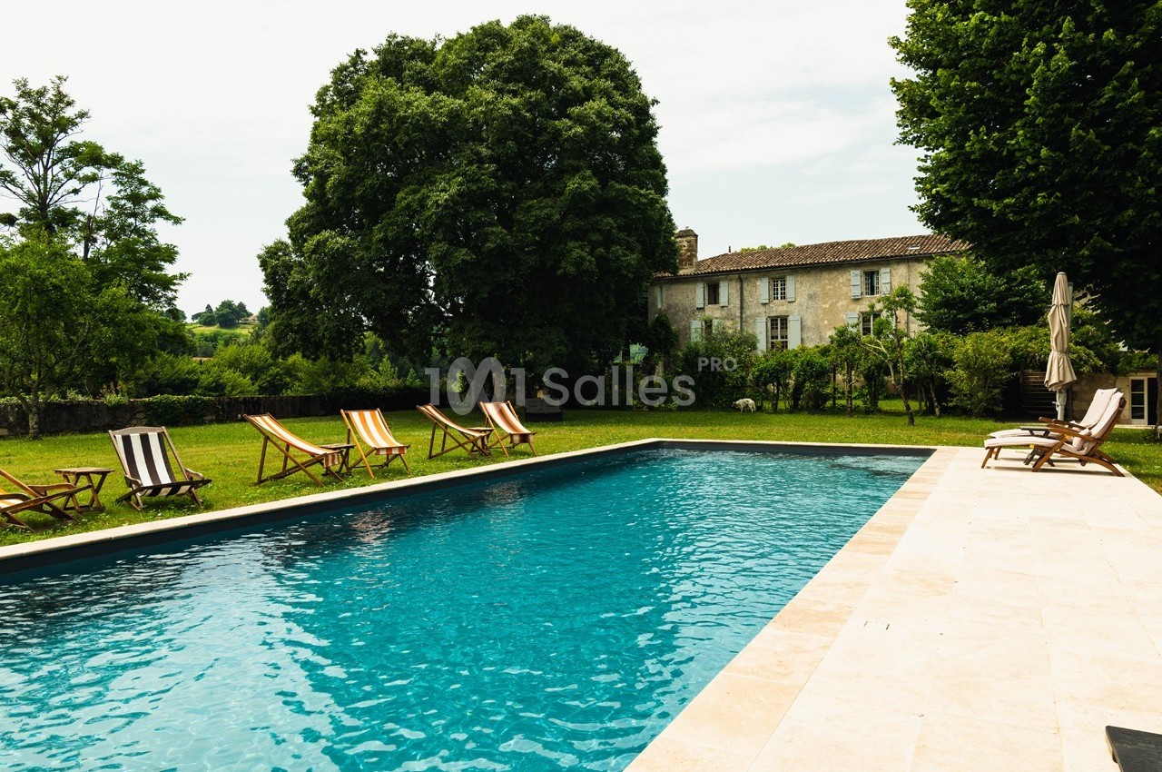 Piscine extérieure entourée de chaises longues, située dans un jardin verdoyant avec une maison en arrière-plan.