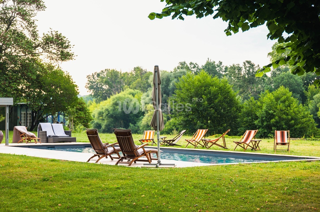 Chaises longues et parasol disposés autour d'une piscine extérieure, entourée de verdure et d'arbres.