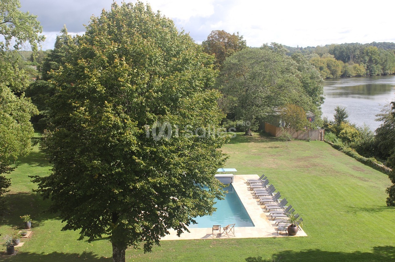 Vue d'un grand jardin verdoyant avec une piscine entourée de chaises longues, près d'une rivière bordée d'arbres.