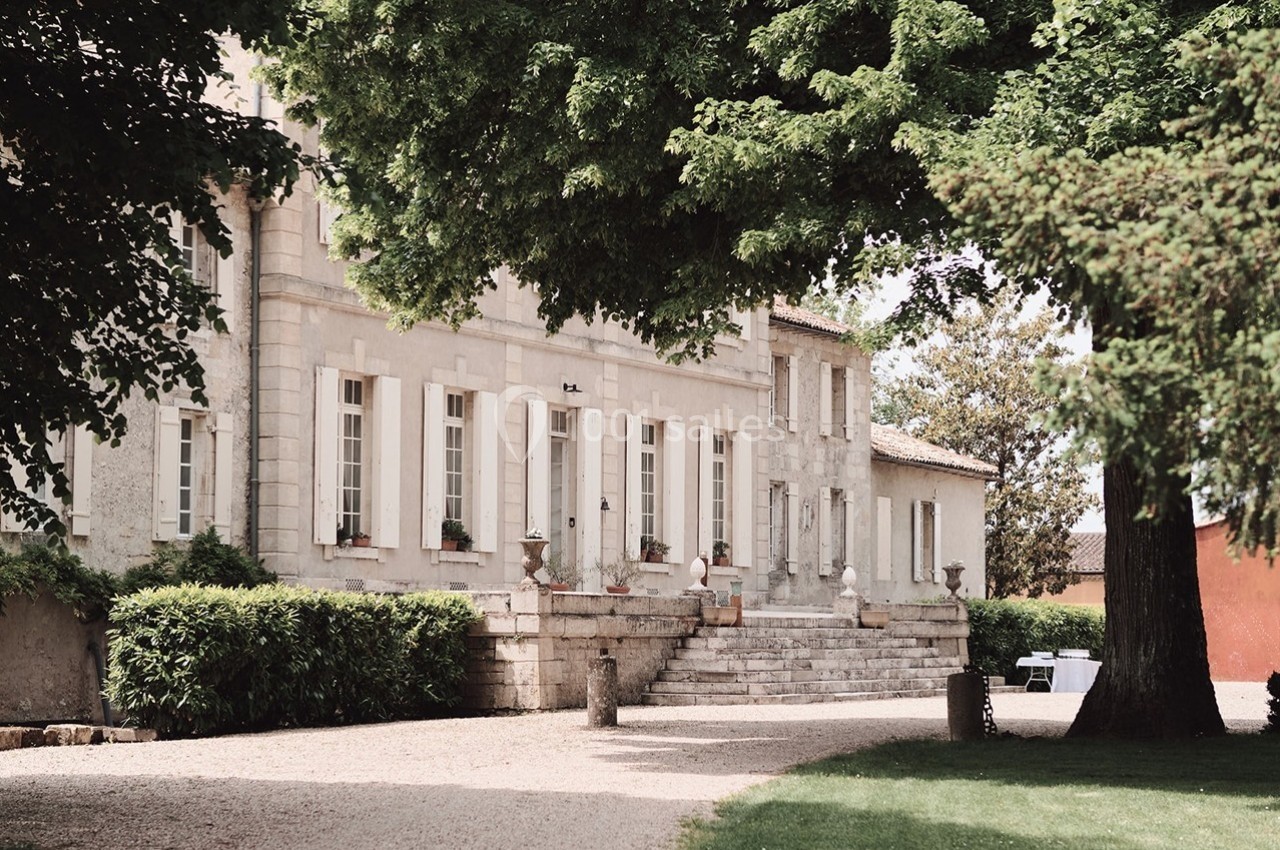 Façade d'une maison en pierre entourée d'arbres, avec un chemin de gravier et des marches menant à l'entrée.
