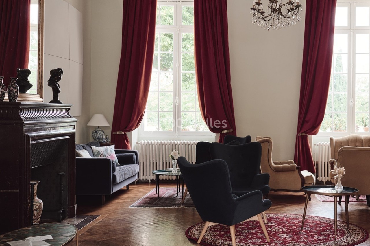 Salon élégant avec fauteuils, canapé, cheminée, lustre et grandes fenêtres encadrées de rideaux rouges.
