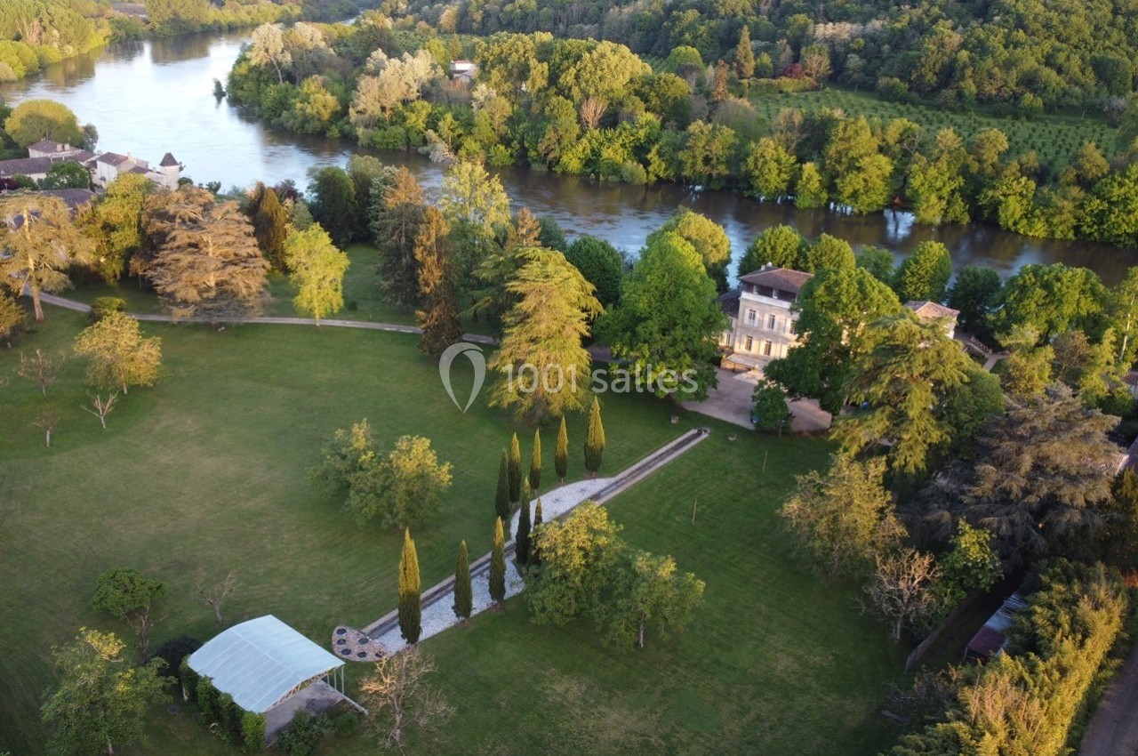 Vue aérienne d'un domaine avec une grande maison, un parc arboré, une rivière et une tente blanche dans un jardin.