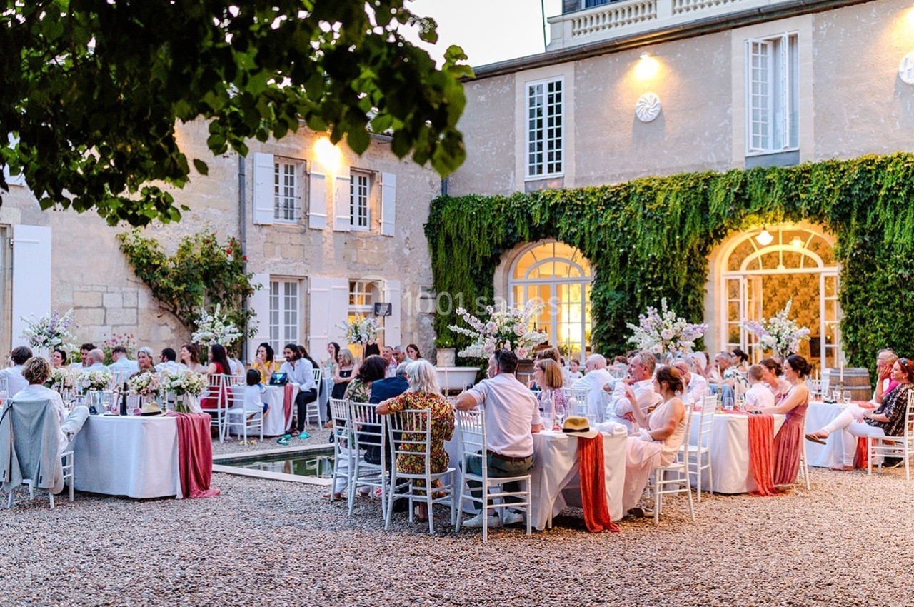 Invités assis à des tables décorées dans la cour d'une maison en pierre, lors d'une réception en plein air.
