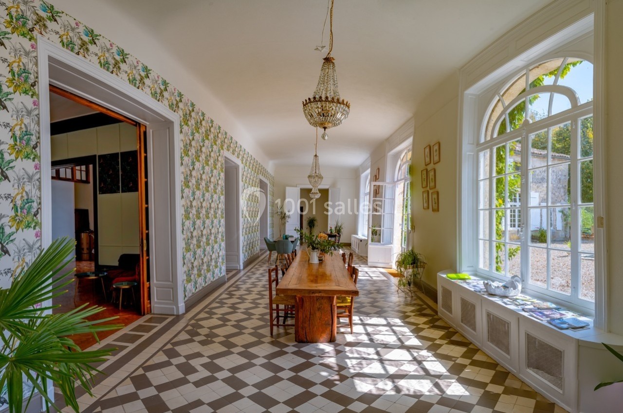 Salle lumineuse avec de grandes fenêtres, table en bois massif, sol en carreaux et murs ornés de papier peint floral.
