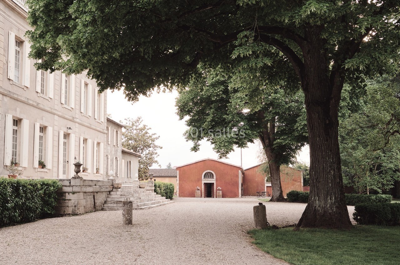 Allée gravillonnée bordée d'arbres menant à un bâtiment rouge, près d'une grande maison en pierre.