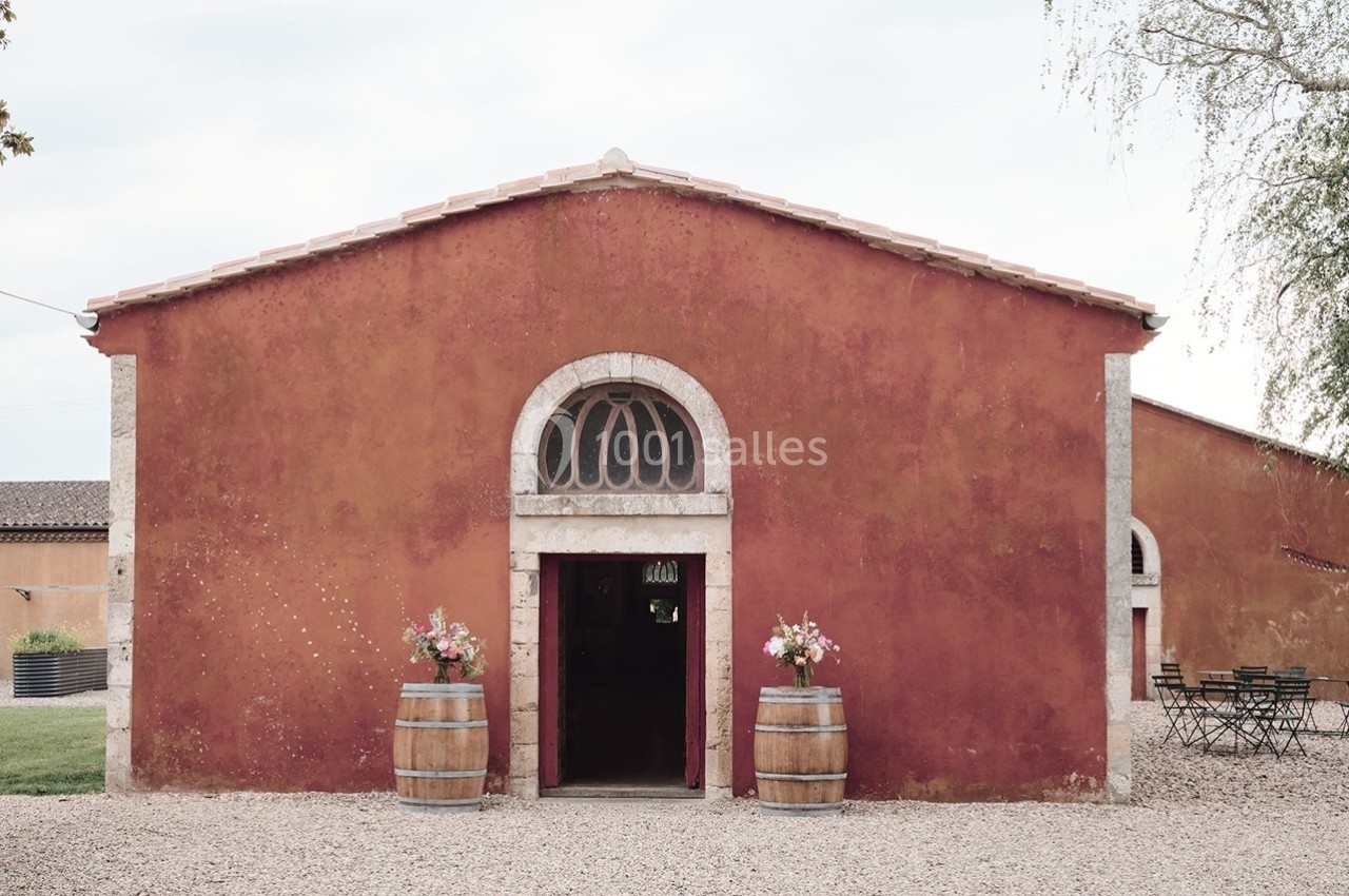 Façade d'un bâtiment en pierre rouge avec une porte voûtée, deux tonneaux décorés de fleurs et une cour gravillonnée.