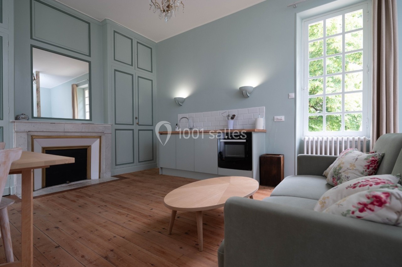 Salon lumineux avec parquet en bois, canapé, table basse, cheminée, cuisine intégrée et grande fenêtre donnant sur l…