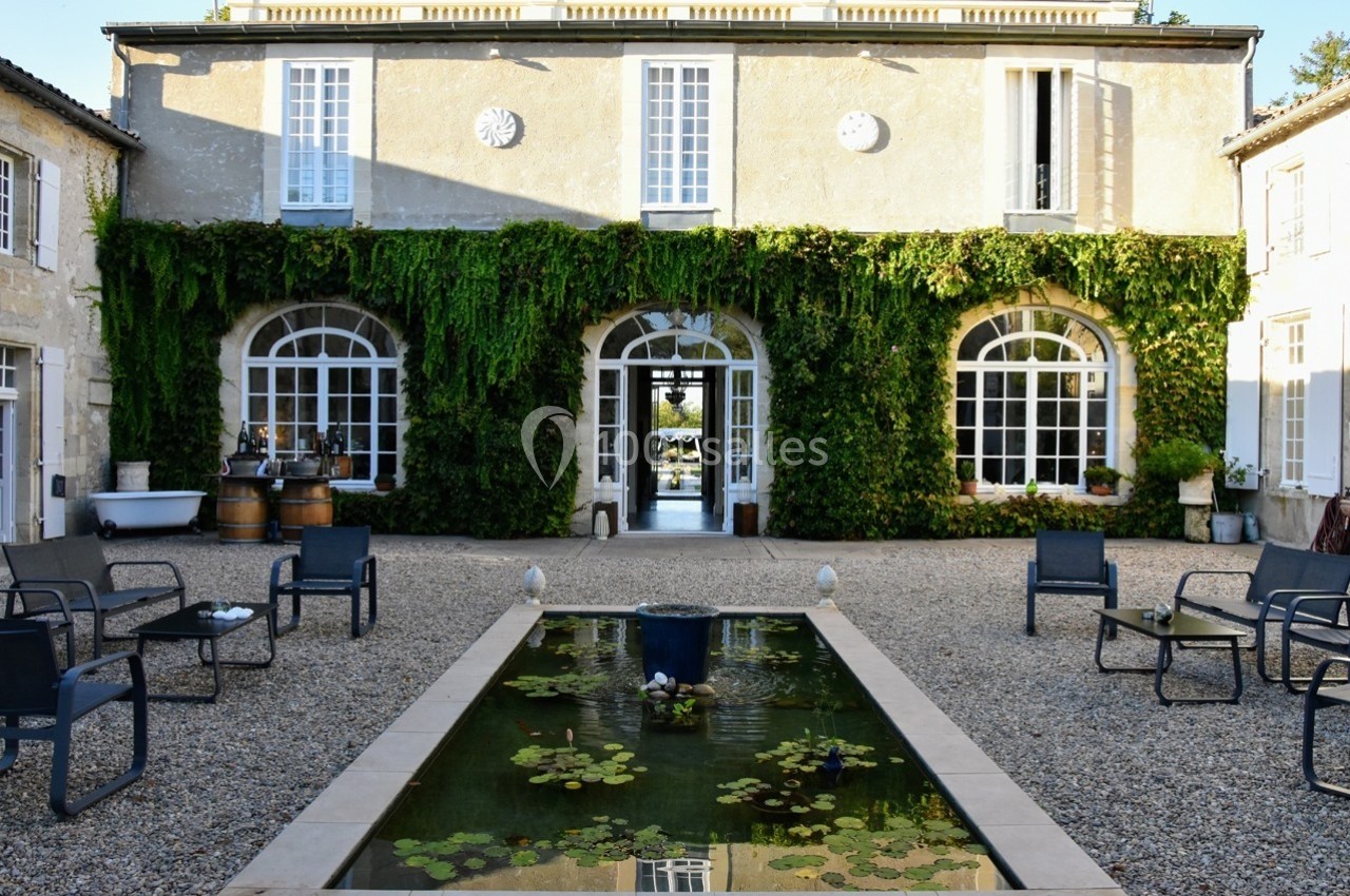Façade d'une maison en pierre recouverte de vigne, avec une cour gravillonnée et un bassin orné de nénuphars.