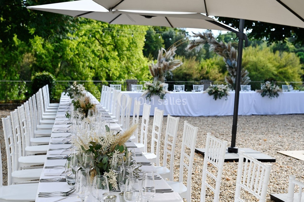 Tables de réception décorées avec des fleurs et alignées en extérieur sous des parasols, entourées de verdure.