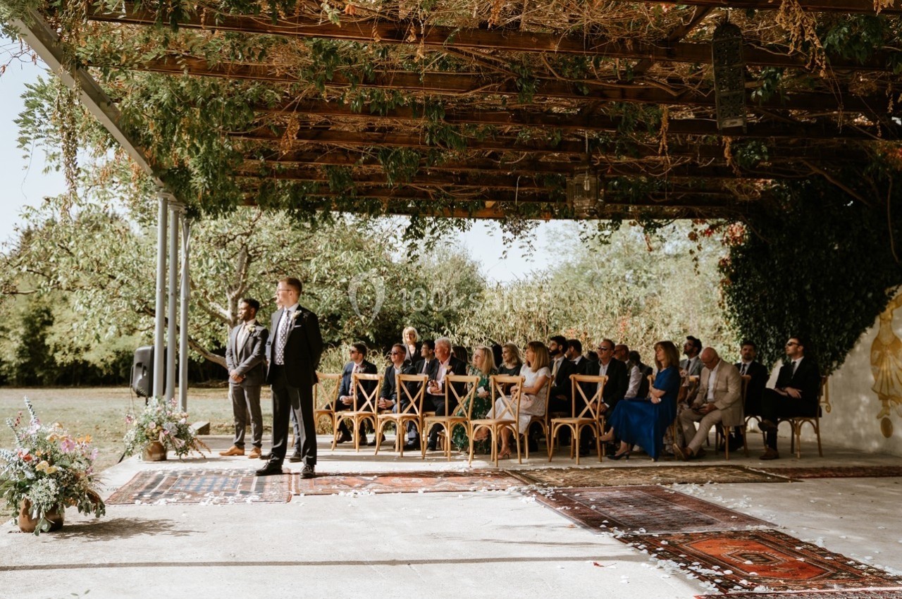 Un mariage en extérieur avec des invités assis sous une pergola ornée de végétation, face à un officiant.