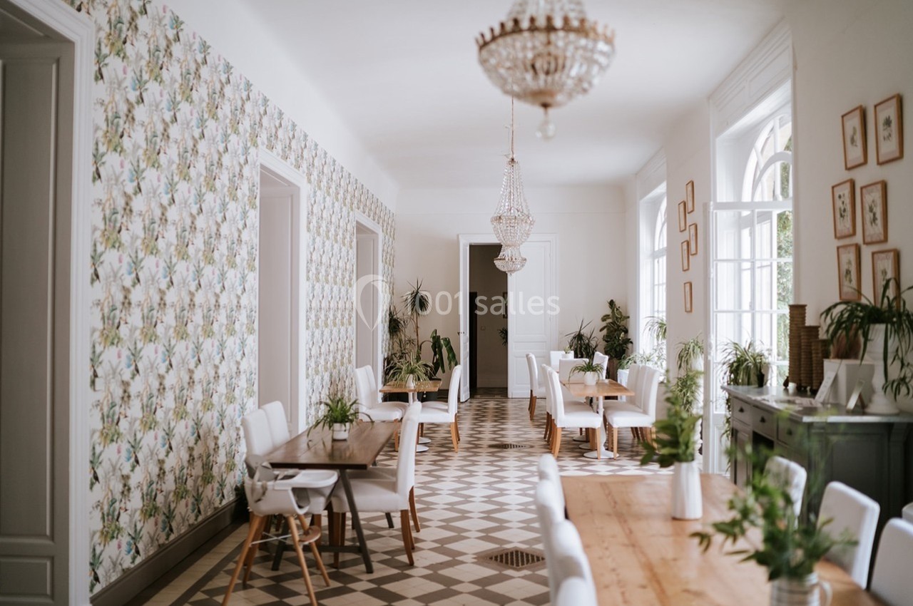Salle lumineuse avec tables en bois, chaises blanches, plantes décoratives et lustres en cristal.
