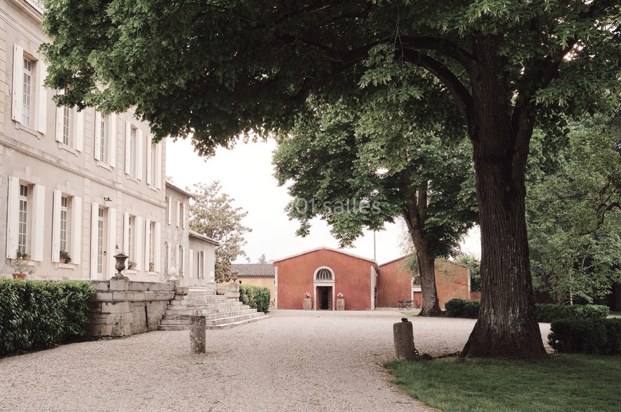 Allée gravillonnée bordée d'arbres menant à une bâtisse en pierre et un bâtiment rouge dans un cadre verdoyant.