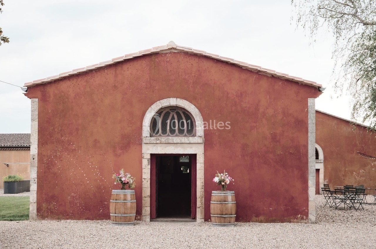 Façade d'un bâtiment en pierre ocre avec une porte voûtée, deux tonneaux fleuris et une cour en gravier.