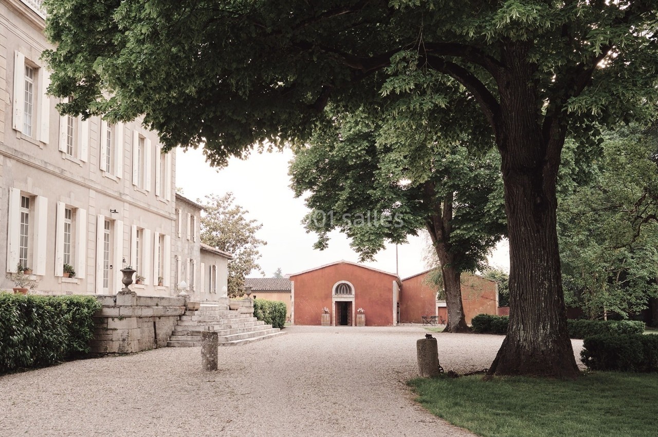 Allée gravillonnée bordée d'arbres menant à un bâtiment en pierre et une dépendance rouge dans un cadre verdoyant.