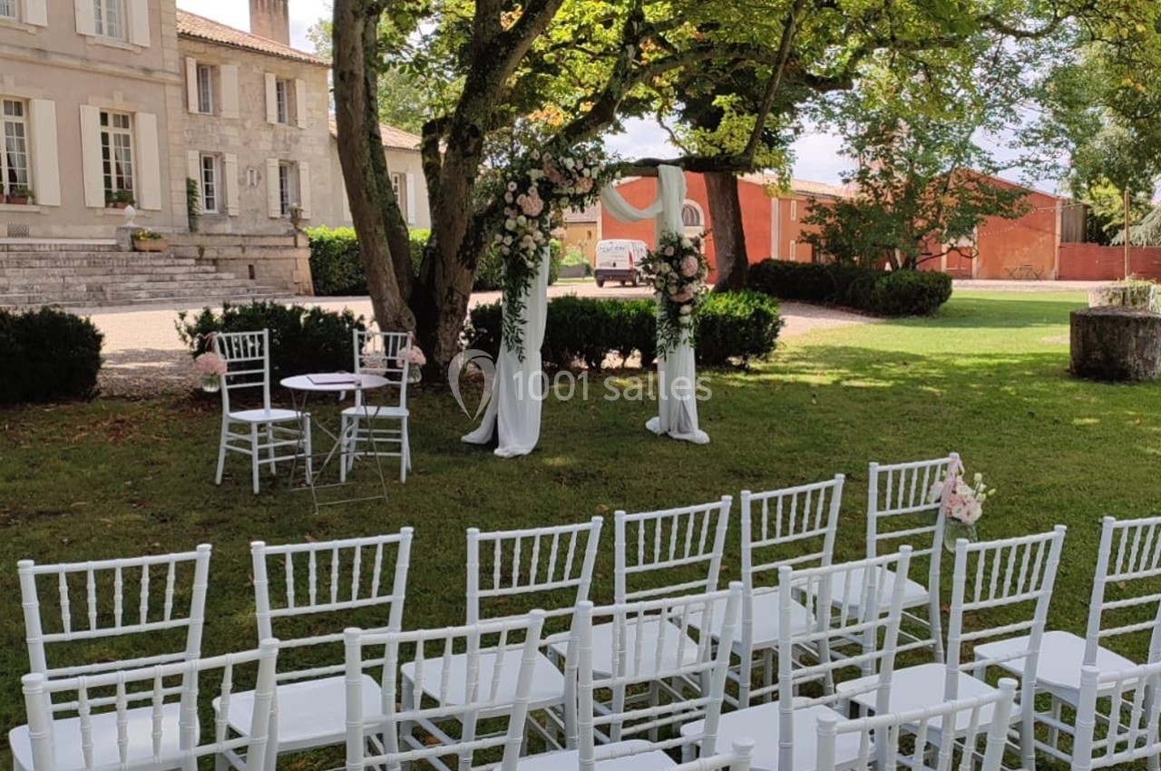 Chaises blanches alignées devant une arche décorée de fleurs dans un jardin, près d'un bâtiment en pierre.