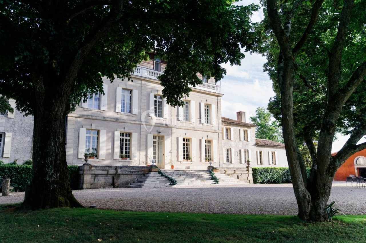 Façade d'une grande maison en pierre entourée d'arbres, avec une allée gravillonnée au premier plan.