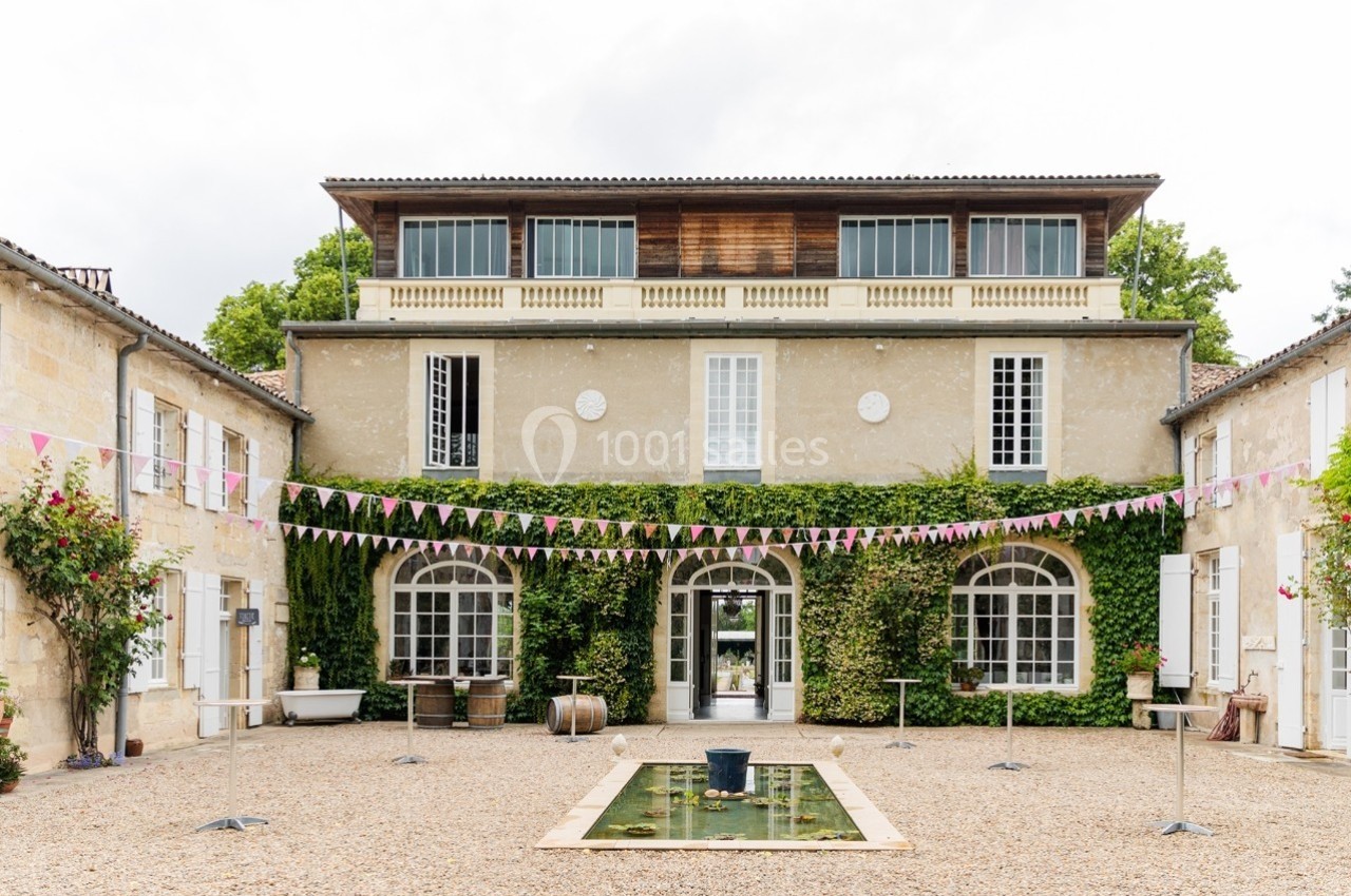Façade d'une grande maison en pierre avec lierre, cour gravillonnée, bassin central et guirlandes suspendues.