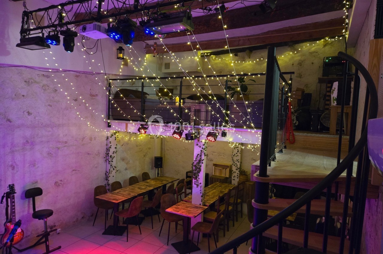 Salle avec tables en bois, guirlandes lumineuses suspendues, mezzanine et escalier en colimaçon.