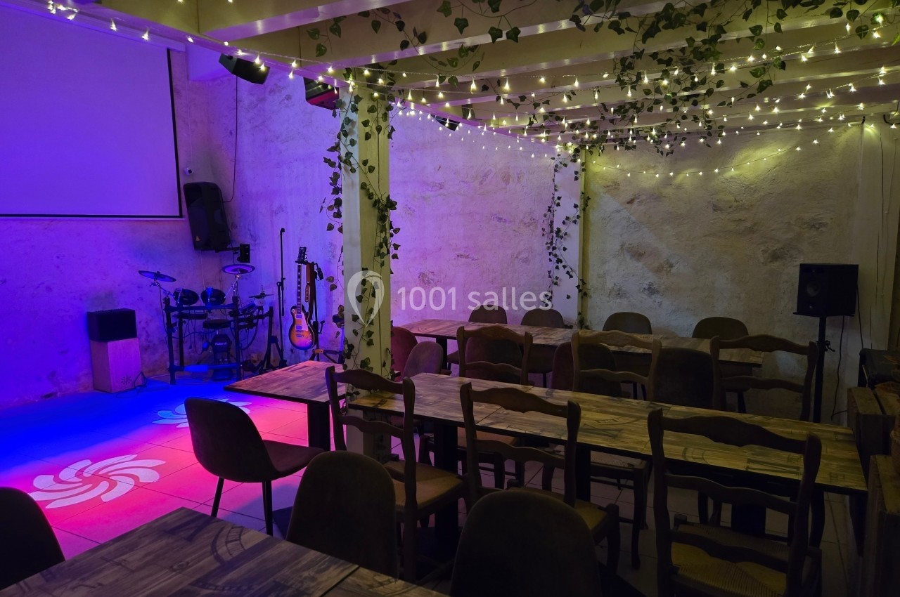 Salle décorée avec des guirlandes lumineuses et des plantes, équipée de tables, chaises et d'une scène avec instruments de…