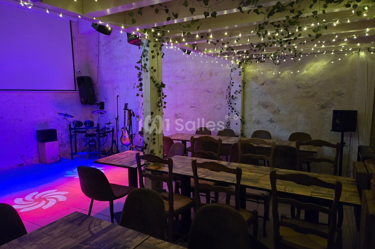 Salle décorée avec des guirlandes lumineuses et des plantes, équipée de tables, chaises et d'une scène avec instruments de…
