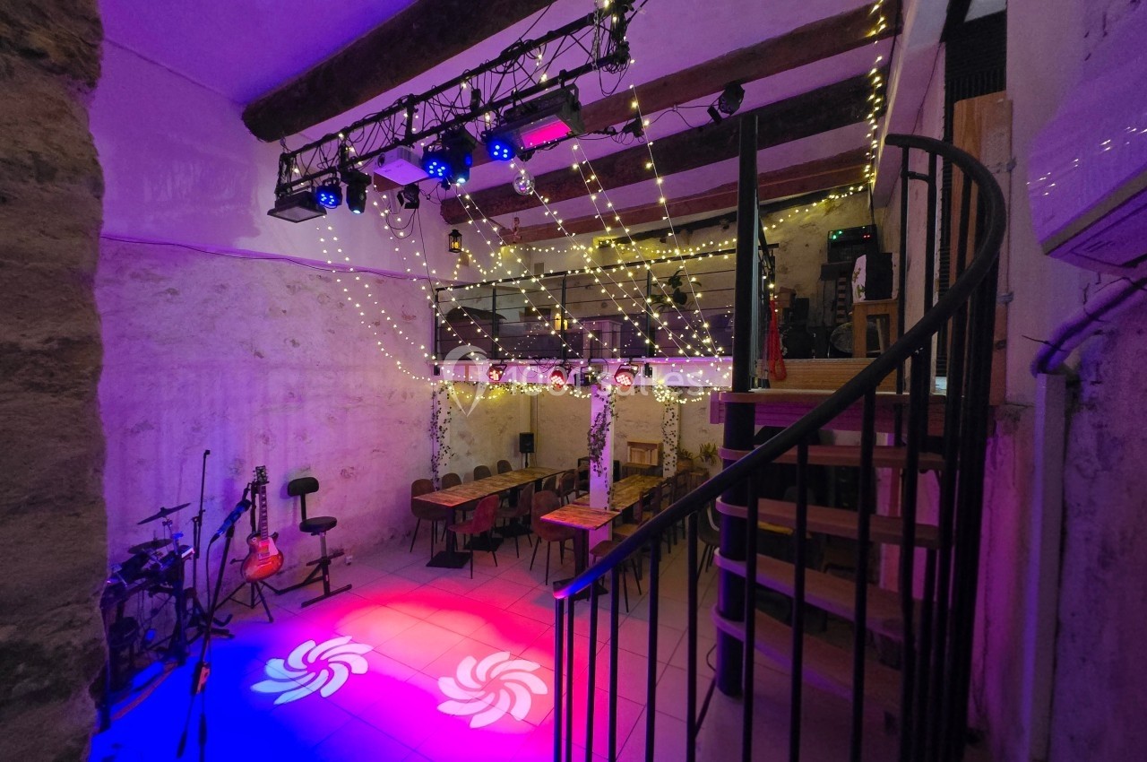 Salle avec éclairage coloré, scène équipée d'instruments de musique, tables et escalier en colimaçon.