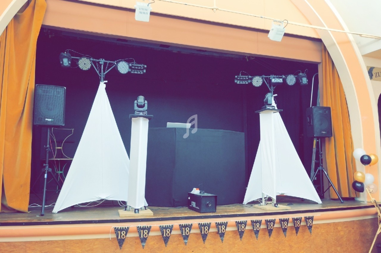 Scène décorée avec des rideaux orange, des enceintes, des éclairages et une table de DJ pour un événement festif.