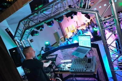 Un DJ mixe sur une scène éclairée, face à une salle avec des invités assis et des lumières colorées.