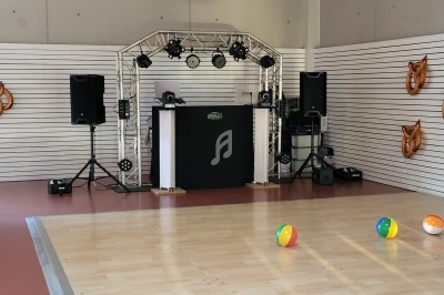 Scène de DJ avec éclairage et enceintes dans une salle, sol en parquet clair, ballons de plage colorés au premier plan.