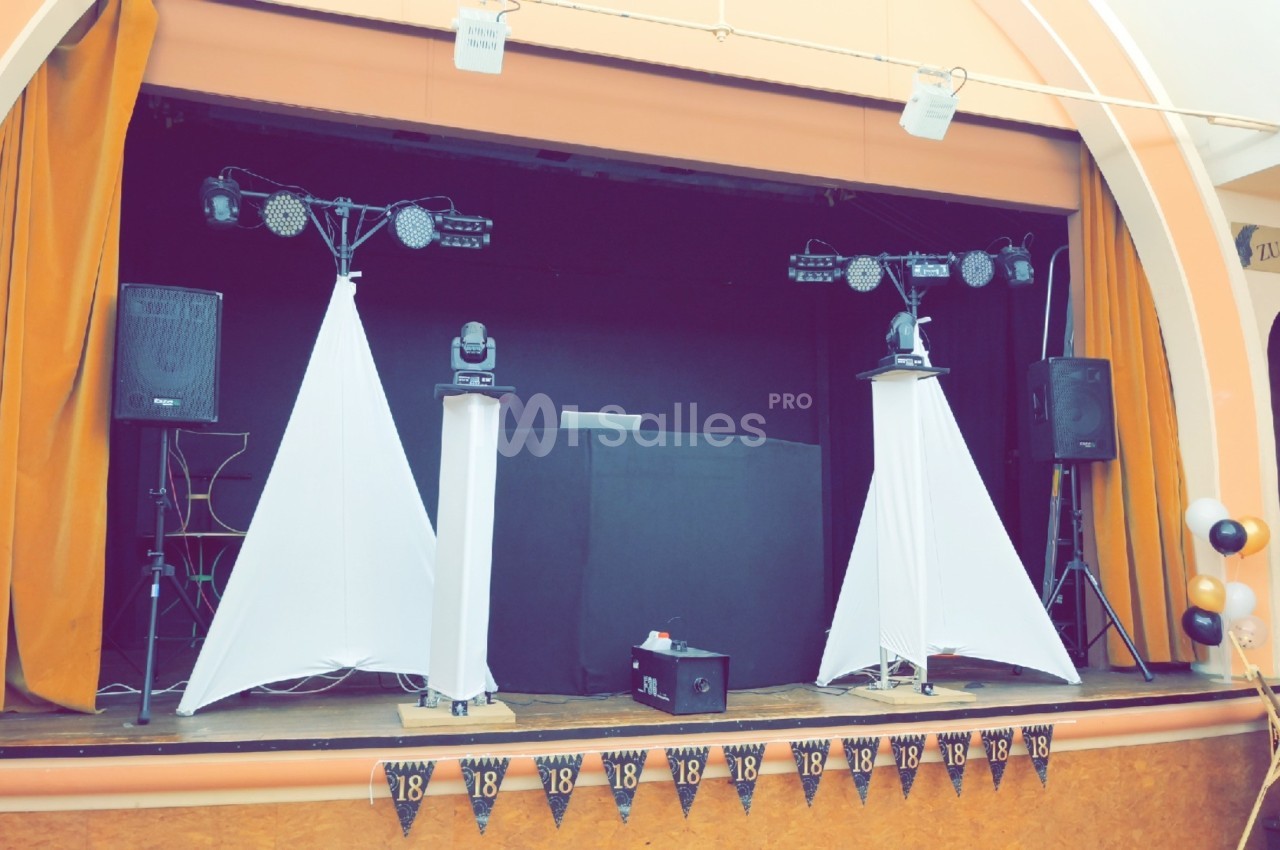 Scène décorée avec des rideaux orange, des enceintes, des lumières et une table de DJ pour un événement festif.