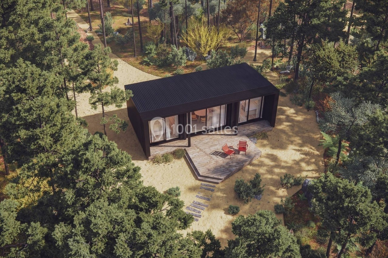Maison moderne en bois sombre entourée de forêt, avec terrasse aménagée et chaises longues sur un sol sablonneux.