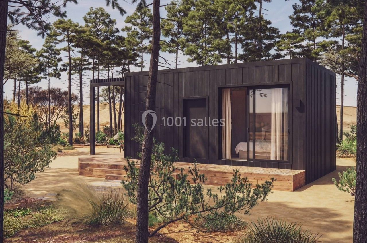 Maison en bois moderne avec terrasse, entourée de végétation et de pins sous un ciel clair.
