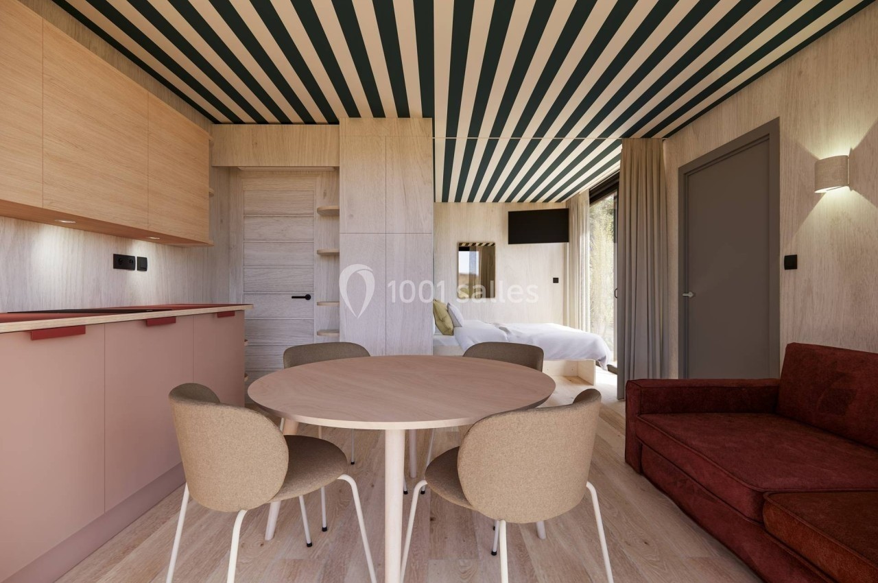 Intérieur d'une pièce lumineuse avec coin cuisine, table ronde, canapé, et lit visible en arrière-plan.