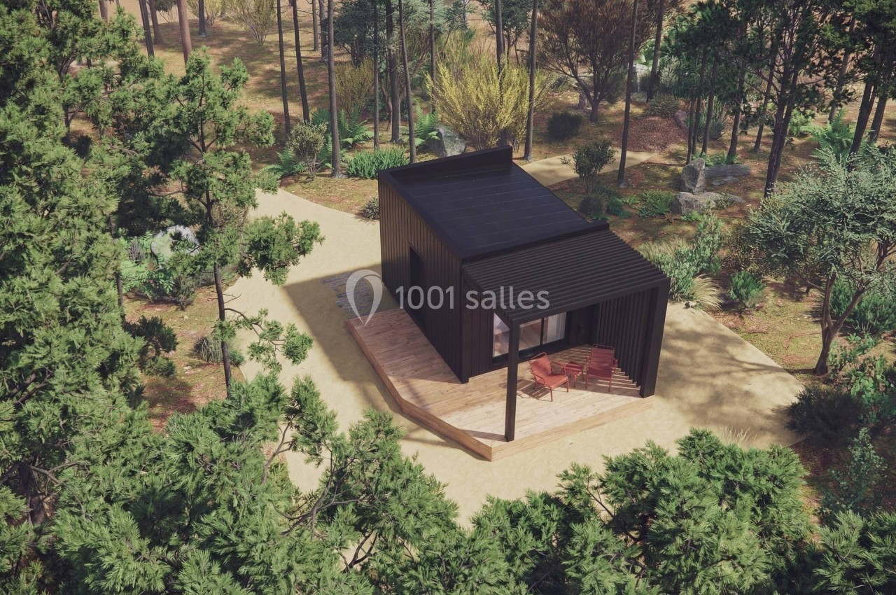 Petite maison moderne en bois noir avec terrasse, située dans une forêt dense et entourée de végétation.