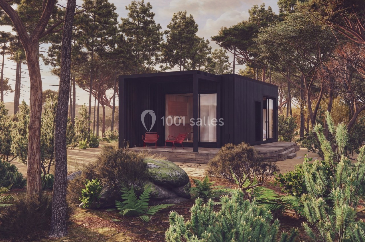 Petite maison moderne noire entourée de végétation dense et de pins, avec deux chaises rouges sur la terrasse.